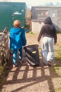 Two children carrying a large black plastic bin together along a muddy path between wooden outbuildings on a sunny day.